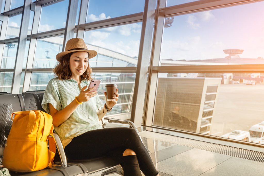 Woman using her phone, sitting in an airport terminal next to a yellow backpack