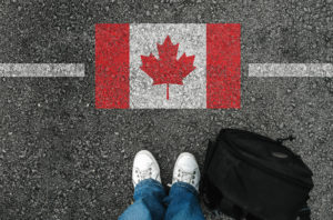 Person in white shoes with a backpack standing at a line in the asphalt marking the Canadian border