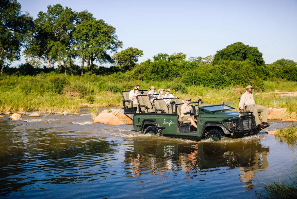 A group of tourists and two tour guides in a safari vehicle driving through a body of water