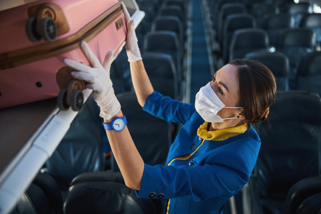 Masked flight attendant putting a suitcase in the overhead bin on an empty plane