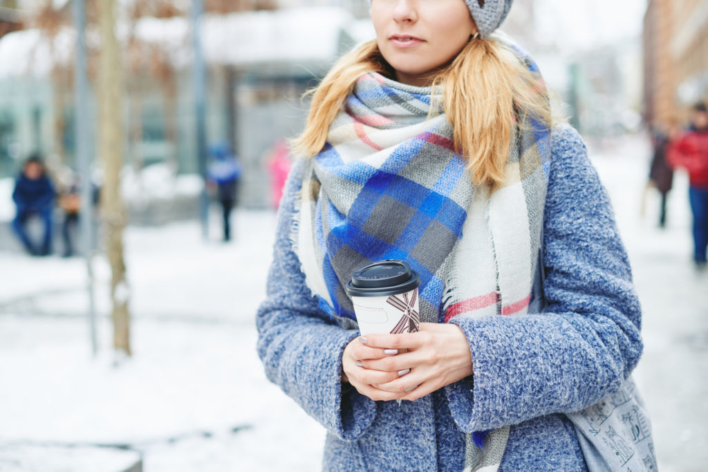 Mid-shot of a woman wearing warm winter clothes and a blanket scarf, holding a travel cup of coffee