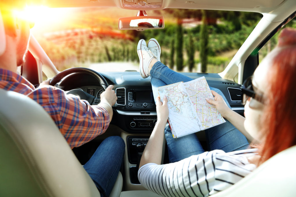 Two people in the front seat of a car, one driving and one with their feet on the dashboard while reading a road trip map