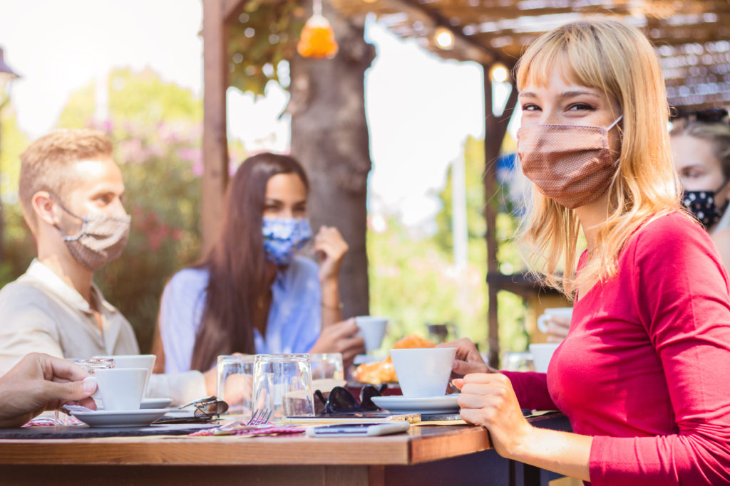 Group of friends at a restaurant table, with focus on blonde woman wearing face mask in foreground