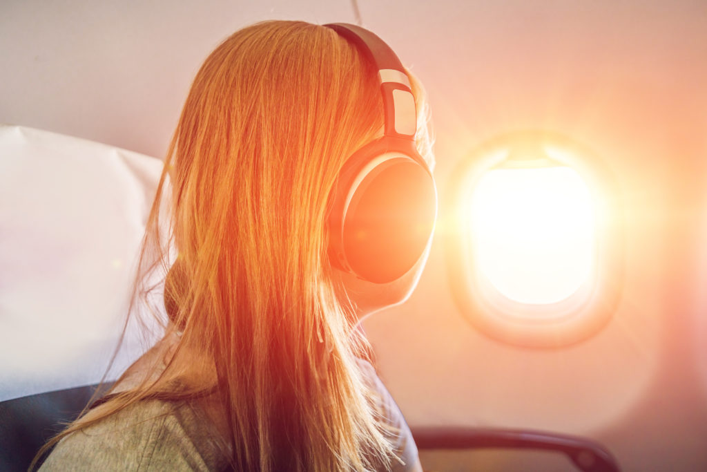 Woman wearing wireless headphones and looking out a plane window at sunset