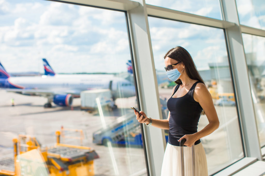 Woman wearing a face mask and checking her phone in front of an airport window with an airplane in the background