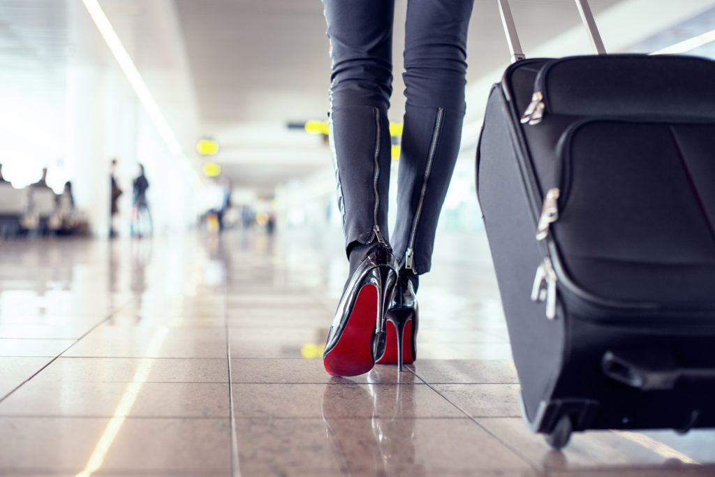 Close up of person's feet at they walk down an airport hallway in high heel shoes while pulling a rolling suitcase